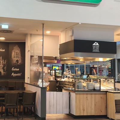 Ein Restaurantbereich mit Tischen, Stühlen, Speisetheke, Wandbildern und Beschriftungen wie „Wir wünschen Guten Appetit“.