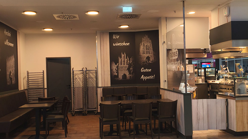 Ein heller Gastraum mit Stühlen und Tischen, Wandbildern mit Schriftzügen, daneben eine offene Küche vom GLOBUS Restaurant.