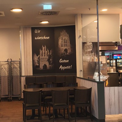 Ein heller Gastraum mit Stühlen und Tischen, Wandbildern mit Schriftzügen, daneben eine offene Küche vom GLOBUS Restaurant.