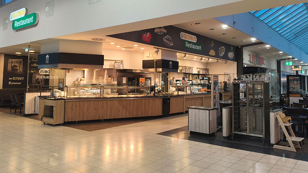 Das GLOBUS Restaurant mit heller Beleuchtung, Holzelementen, Ausgabetheke und Sitzbereich in einer Einkaufszentrumspassage.