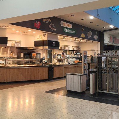 Das GLOBUS Restaurant mit heller Beleuchtung, Holzelementen, Ausgabetheke und Sitzbereich in einer Einkaufszentrumspassage.