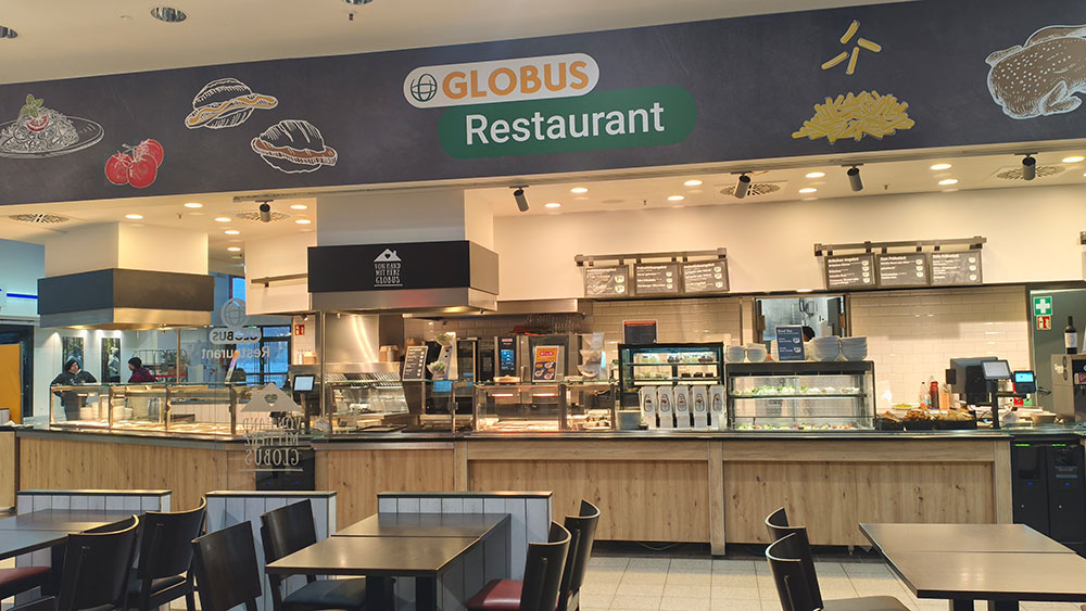 Theke mit Speisen hinter Glas im GLOBUS Restaurant unter einem Schild mit dem GLOBUS Logo und Speiseillustrationen.