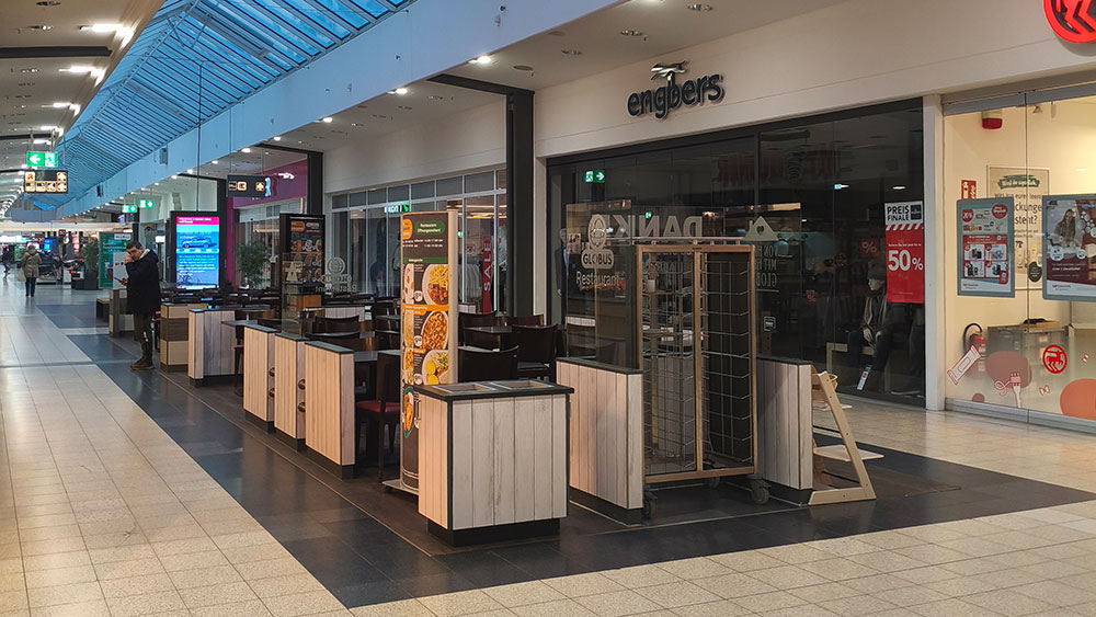 Sitzbereich des GLOBUS Restaurants in einer Einkaufspassage, daneben Geschäft „engbers“ und Werbetafel mit Rabattaktionen.