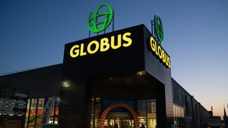 Der GLOBUS Eingang bei Abenddämmerung mit beleuchtetem Logo, einem Torbogen aus Luftballons und Kunden mit Einkaufswagen.