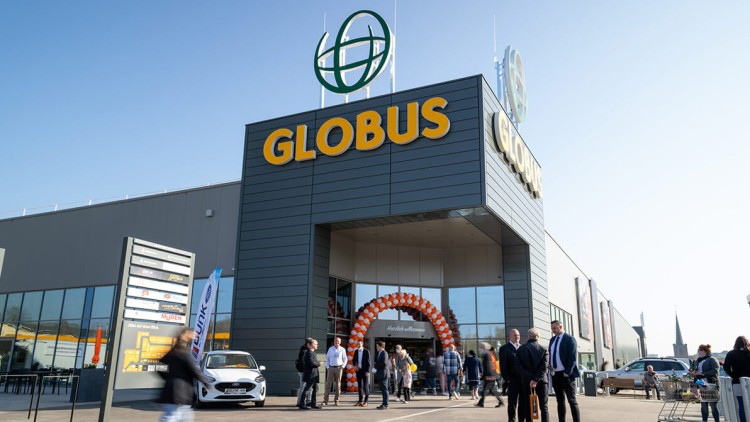 Eingang eines GLOBUS Marktes mit orangenen und weißen Luftballons, Besuchende gehen hinein oder stehen davor im Gespräch.