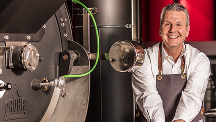 Eine Person in Schürze hält geröstete Kaffeebohnen in den Händen vor einer großen Kaffeeröstmaschine.