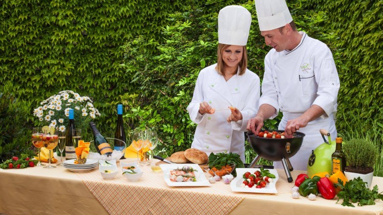 Zwei Köche in weißen Jacken bereiten eine Mahlzeit an einem dekorierten Tisch mit Gemüse, Wein und Blumen vor.