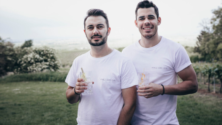 Zwei Personen in weißen T-Shirts mit Aufschrift „PFALZ DRY GIN“ halten Gin-Gläser, im Hintergrund eine grüne Landschaft.
