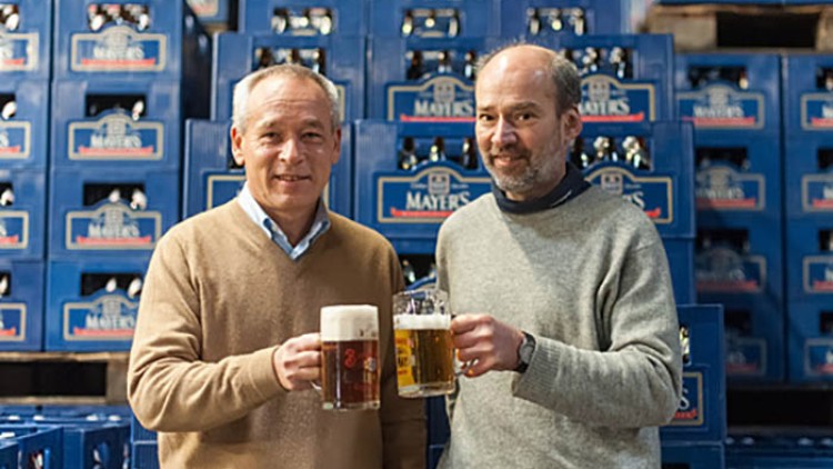 Zwei Personen halten Biergläser vor Kistenstapeln mit der Aufschrift Mayers, im Hintergrund blaue Getränkekisten.