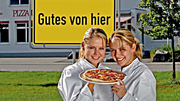 Zwei Personen in weißen Jacken halten eine Pizza vor einem gelben Schild mit der Aufschrift Gutes von hier.