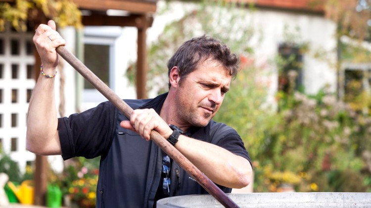 Person rührt mit einem langen Holzstab in einem großen silberfarbenen Behälter, im Hintergrund ein Garten mit Bäumen und Haus.