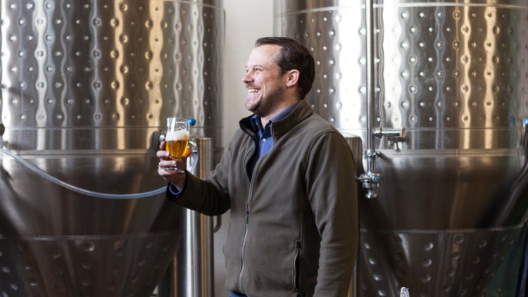 Person mit Glas Bier steht vor großen Edelstahl-Braukesseln und umgebenden Schläuchen in einer Brauerei.