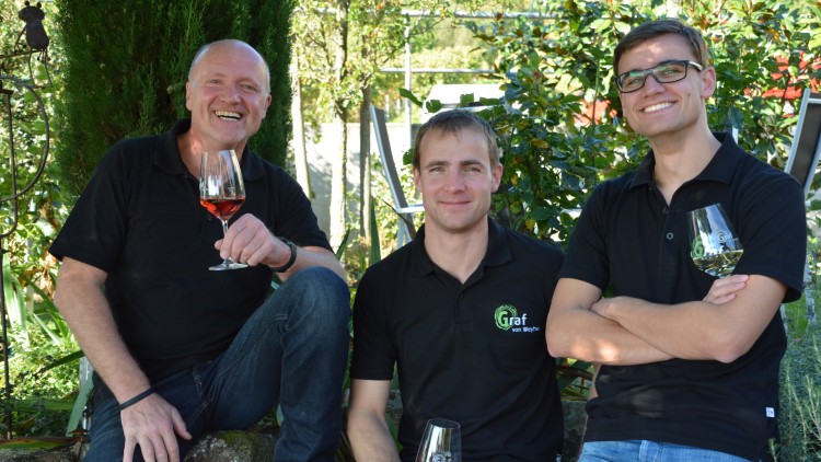 Drei Personen in schwarzen Polo-Shirts mit Graf-Logo, halten Gläser mit Wein, umgeben von Pflanzen im Hintergrund.