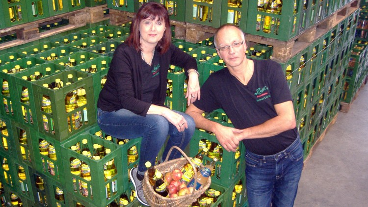 Person sitzt auf grünen Getränkekisten, daneben steht eine Person, vorne steht ein Korb mit Äpfeln und einer Flasche.