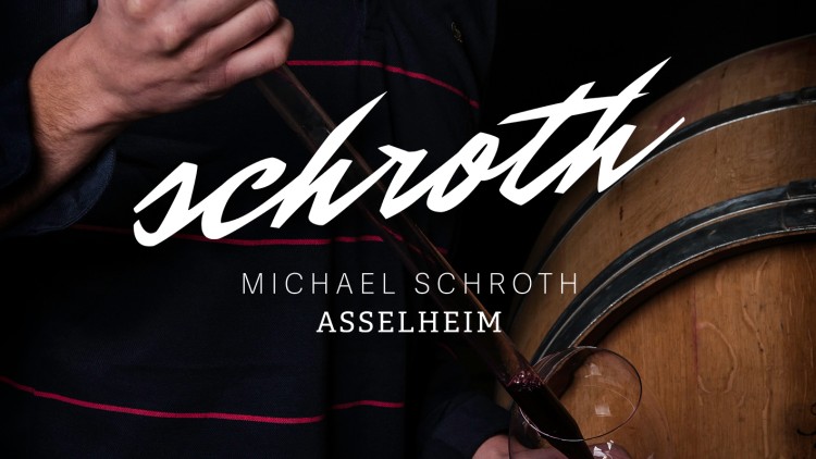 Person in dunkel gestreiftem Oberteil hält ein Weinglas vor einem Holzfass, Schriftzug „Schroth Michael Schroth Asselheim“ ist sichtbar.