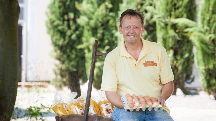 Person in gelbem Hemd mit Logo hält ein Tablett mit Eiern, daneben ein Holzwagen mit verpackten Nudeln, Bäume im Hintergrund.