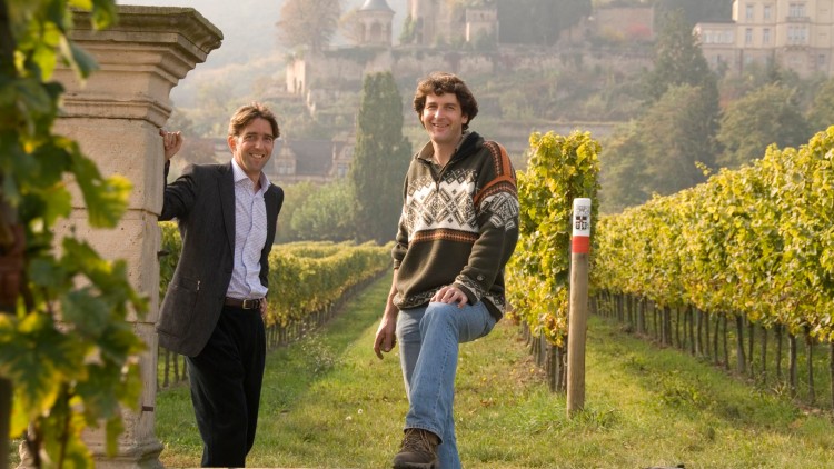 Zwei Personen stehen in einem Weinberg mit Schloss im Hintergrund, ein Steinpfeiler und ein Holzpfosten mit Markierung sind sichtbar.