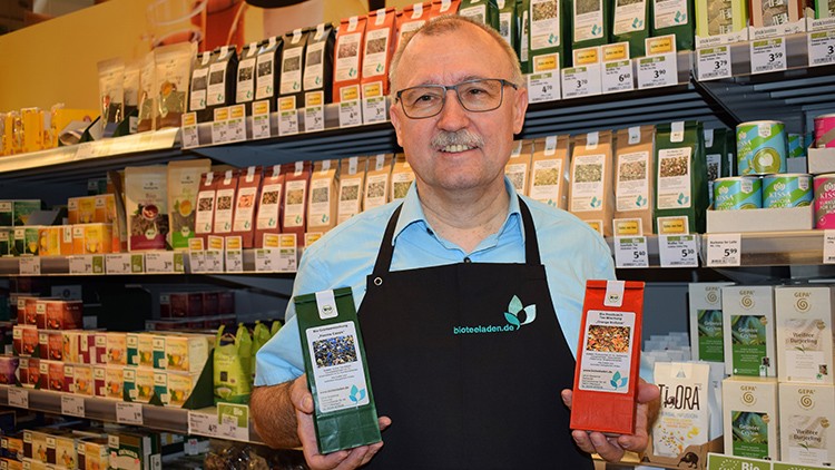Eine Person in Schürze hält zwei Packungen Tee vor einem Regal mit verschiedenen Teeprodukten, oben ist „bioteeladen.de“ zu lesen.