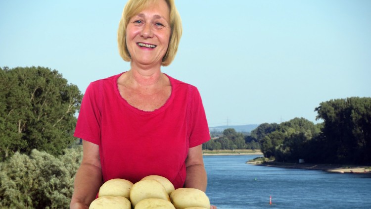 Eine Person in rotem T-Shirt hält einen Holzteller mit mehreren hellen Brötchen, im Hintergrund ist ein Fluss und Wald zu sehen.