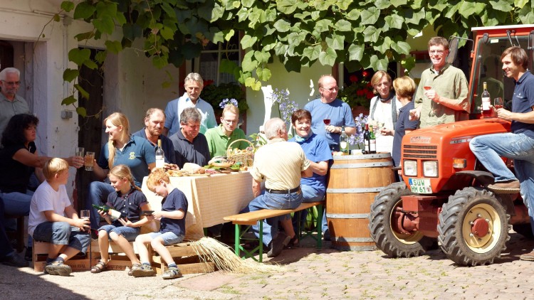 Menschen sitzen und stehen an einem gedeckten Tisch mit Speisen und Getränken im Innenhof, neben einem roten Traktor.