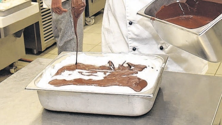 Koch in weißer Uniform gießt flüssige Schokolade aus einer Metallschüssel auf ein Blech mit weißer Masse in einer Küche.