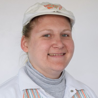 Person trägt eine weiße GLOBUS-Mütze, ein grau-weißes Oberteil und eine Schürze mit orange-grün-weißen Streifen.