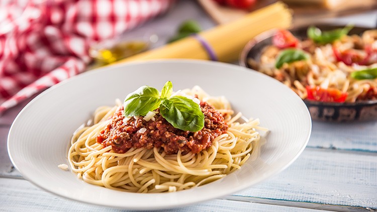 Ein weißer Teller mit Spaghetti, Tomatensauce, geriebenem Käse und frischem Basilikum, im Hintergrund weitere Zutaten.
