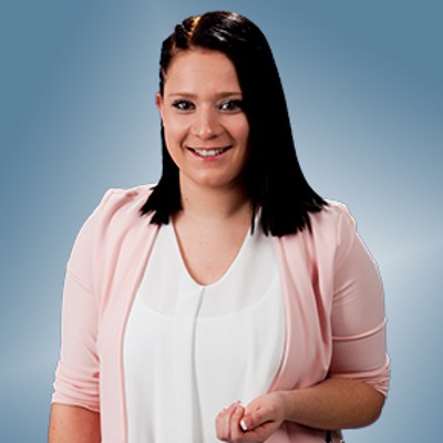 Person trägt eine weiße, fließende Bluse und einen rosafarbenen Blazer vor einem blauen Hintergrund.