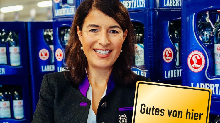 Person in traditioneller Tracht vor blauen Getränkekisten, gelbes Schild mit schwarzer Aufschrift „Gutes von hier“.