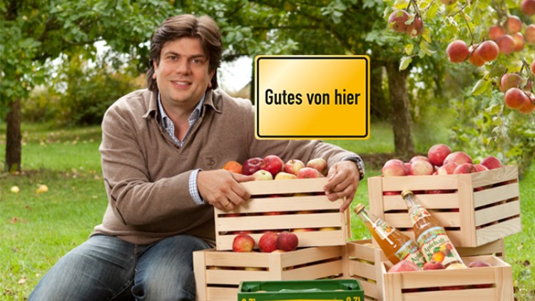 Holzkisten mit roten Äpfeln und Getränkeflaschen stehen auf einer Wiese, gelbes Schild mit Text „Gutes von hier“ im Hintergrund.