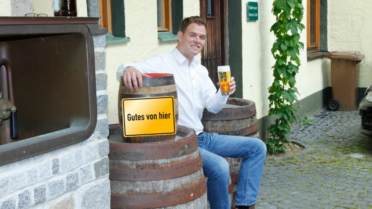 Mann in weißem Hemd sitzt auf Holzfässern vor gelber Wand, hält ein Bierglas, Schild mit „Gutes von hier“ sichtbar.