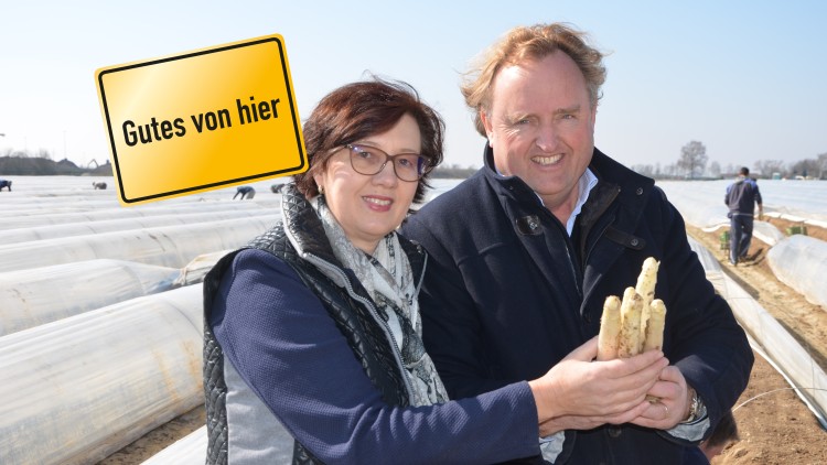 Zwei Personen halten weißen Spargel vor einem Feld mit Spargelreihen, gelbes Schild mit Text Gutes von hier im Bild.