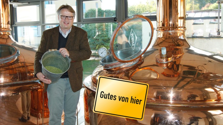 Person hält einen Eimer mit grünem Hopfen vor glänzenden Kupferkesseln, im Vordergrund ein Schild mit der Aufschrift Gutes von hier.