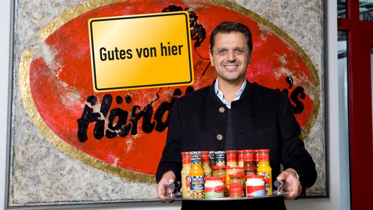 Ein Tablett mit verschiedenen Händlmaier-Produkten steht vor einer Wand mit rotem Händlmaier-Logo und dem Schild „Gutes von hier“.