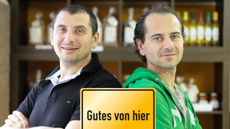Zwei Personen stehen mit verschränkten Armen vor Regalen, gelbes Schild mit der Aufschrift Gutes von hier im Vordergrund.