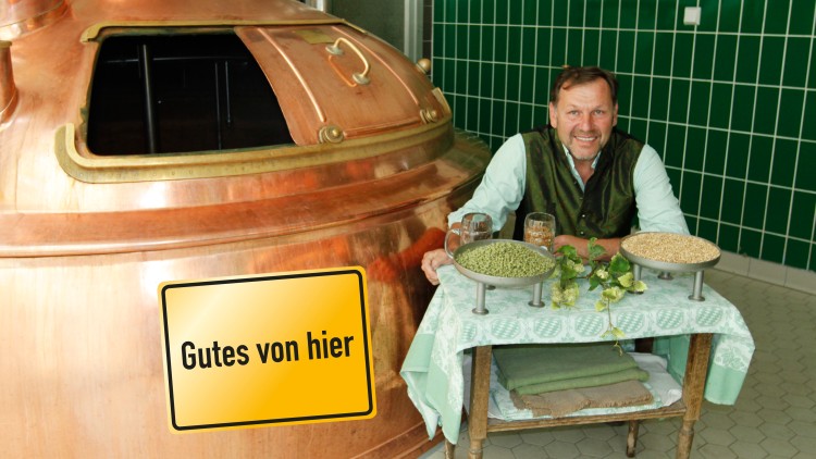 Kupfernes Sudhaus mit geöffneter Klappe, Schild mit „Gutes von hier“ und Tisch mit Hopfen, Malz, Gläsern und Dekoration.