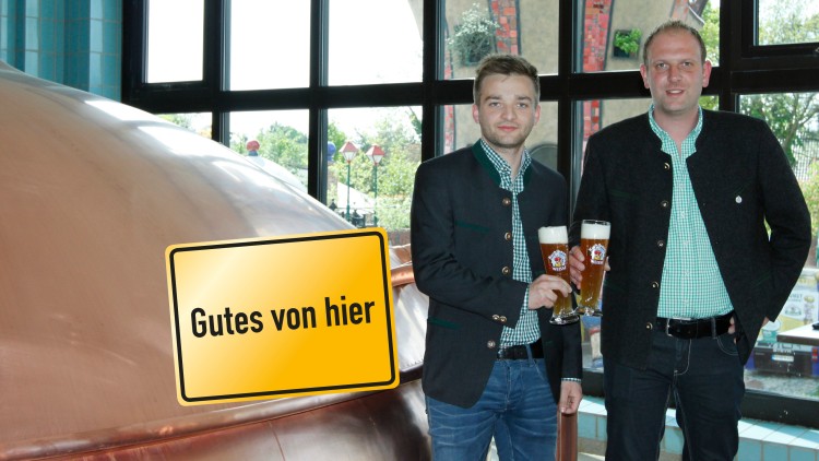 Zwei Personen in Trachten halten Gläser mit Bier vor einem Kupferkessel, daneben ein Schild mit der Aufschrift Gutes von hier.