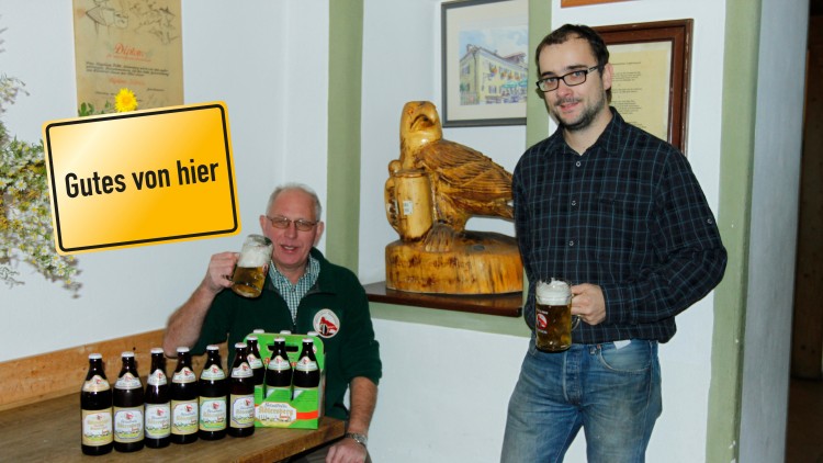 Mehrere Flaschen Bier und ein Getränkekarton stehen auf einem Holztisch, Schild mit Aufschrift Gutes von hier und Holzfigur im Hintergrund.