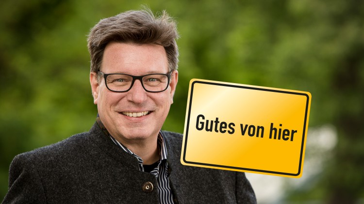 Person in grauer Tracht mit gelbem Schild mit schwarzem Rand und der Aufschrift Gutes von hier, unscharfer grüner Hintergrund.