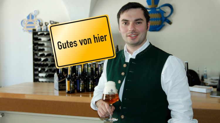 Mann in traditioneller Kleidung hält ein Bierglas, im Hintergrund ein Tresen mit Bierflaschen und das Schild „Gutes von hier“.