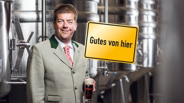 Person im hellgrünen Trachtenanzug hält ein Bierglas mit „Stöttner“-Aufschrift, im Hintergrund metallische Braukessel, Schild „Gutes von hier“.