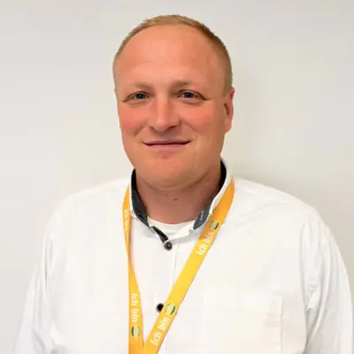 Ein lächelnder Mensch in weißem Hemd mit ID-Band um den Hals vor weißem Hintergrund.