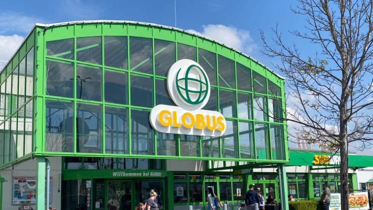 Ein GLOBUS Markt mit grünem Glasbau, Logo über dem Eingang, Menschen und Büschen davor, blauer Himmel mit Wolken oben.