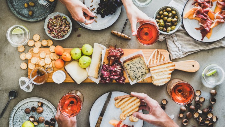 Mehrere gedeckte Teller mit Aufschnitt, Nüssen, Crackern, Käse, Obst und Gläsern mit Getränken auf einem Tisch.