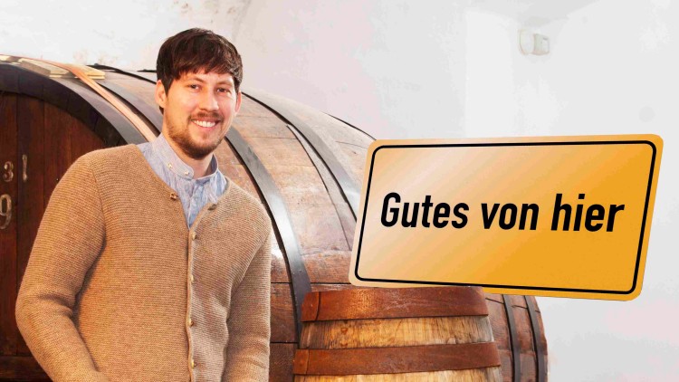 Mann in brauner Strickjacke zapft Bier aus einem Holzfass, daneben stehen mehrere Bierflaschen auf einem Holztisch, Schild \