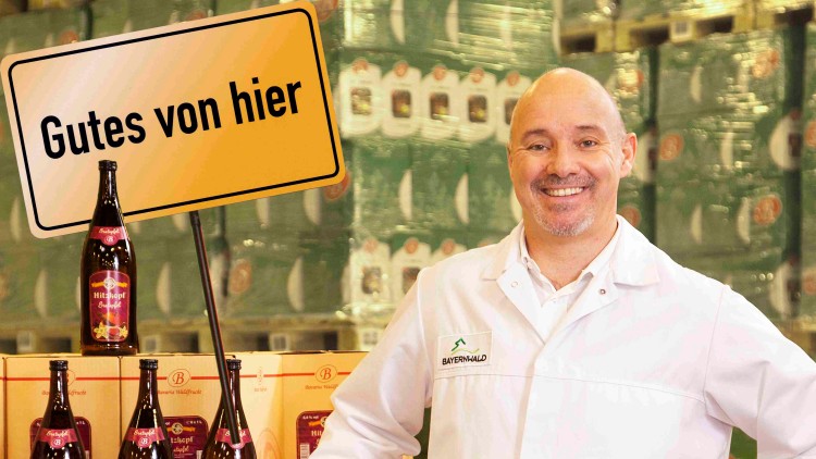 Ein Mann in weißem Arbeitskittel steht neben gestapelten Getränkekartons und Flaschen, ein Schild mit \