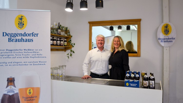 Theke mit Deggendorfer Brauhaus-Logo, Bierflaschen, Gläsern, Personen und Spiegel im hellen Raum mit weißen Wänden.