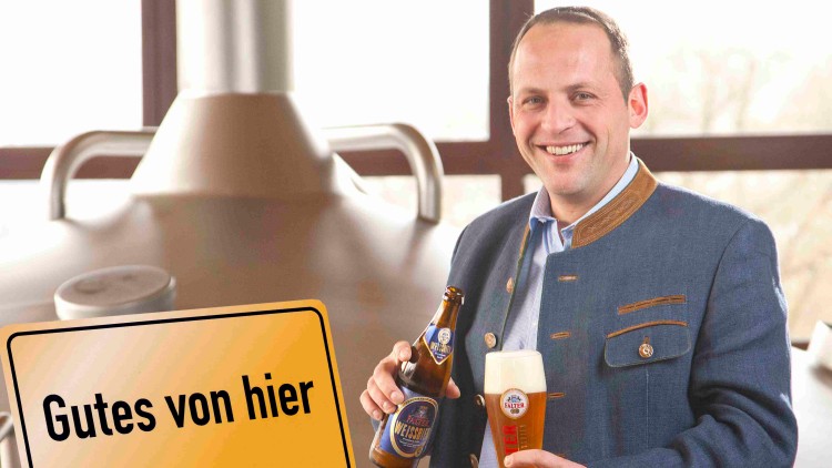 Mann in Trachtenjacke hält eine Flasche Helles Weissbier und ein gefülltes Bierglas vor Sudkesseln, Schild mit „Gutes von hier“.