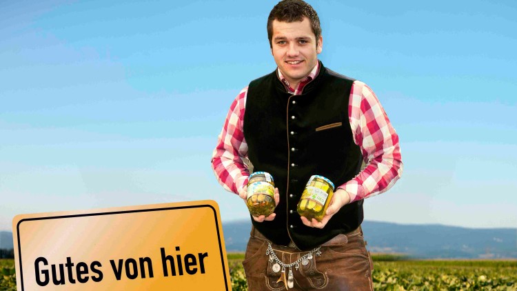 Person in Tracht steht auf einem Feld mit zwei Gläsern Gurken in den Händen, daneben Schild mit Aufschrift Gutes von hier.