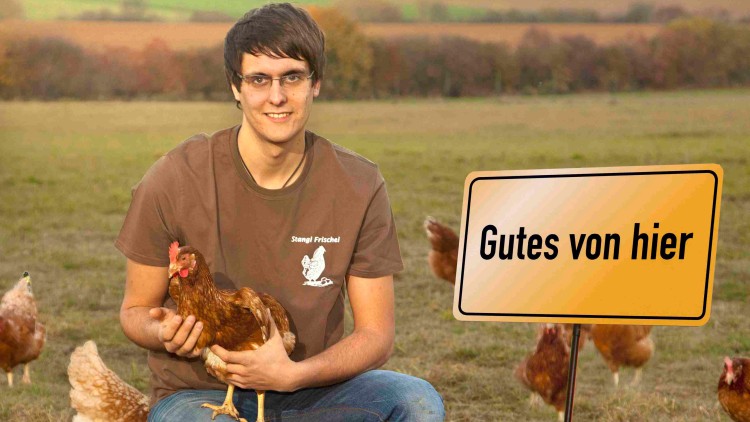 Person in braunem Shirt mit Aufschrift „Snagl Frischei“ hält ein Huhn, im Hintergrund Hühner und ein Schild mit „Gutes von hier“.